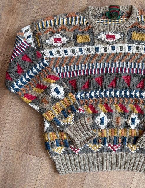 A patterned knit sweater in grey, red, yellow and blue, laid flat on a wood floor